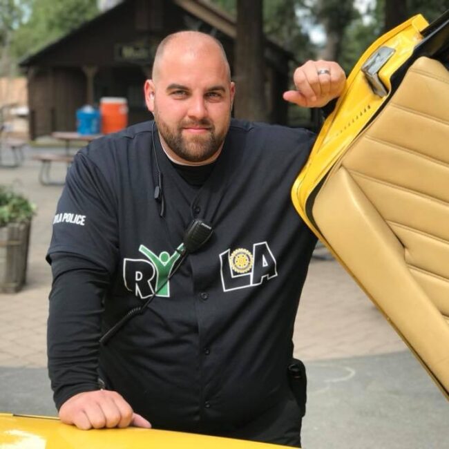 Man in a black jersey with a police badge, leaning on a yellow car door, surrounded by trees and outdoor seating areas, representing community engagement and service.